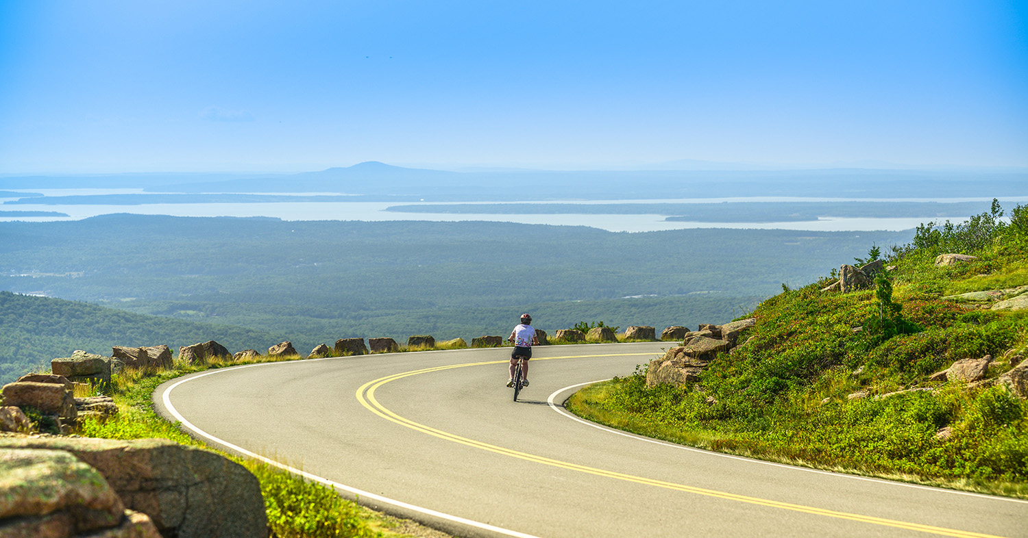 Female mountain bike cyclist riding downhill along Cadillac Mountain road in Acadia National Park, a clear summer day with panoramic view of Acadia National Park (Maine, USA) landscape with lakes.